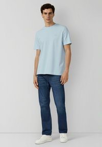 Hellblaues T-Shirt mit rundem Ausschnitt, kurzen Ärmeln und lockerem Schnitt, kombiniert mit dunkelblauen Jeans und weißen Turnschuhen.