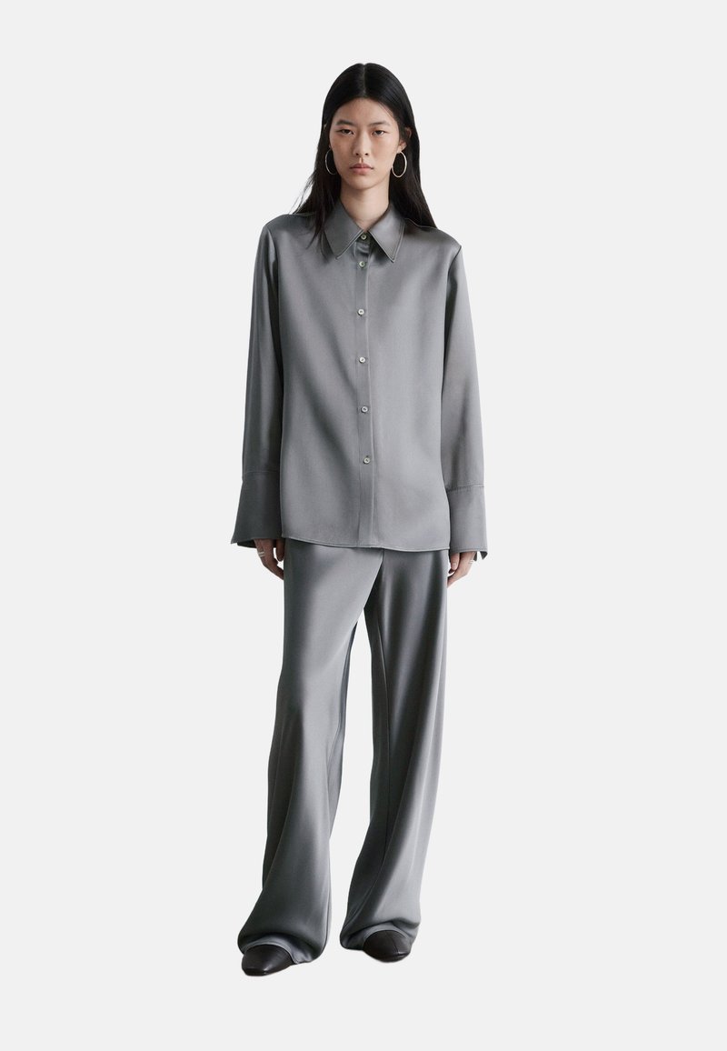 Chemise grise en satin à boutons avec col pointu et manches longues, assortie à un pantalon large de la même matière. Texture lisse, design raffiné.