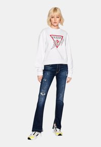 Sweatshirt blanc avec un logo triangulaire rouge, associé à un jean déchiré bleu foncé. Les baskets ont des accents jaunes et des détails noirs et blancs.