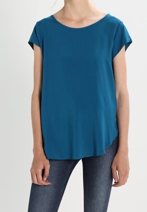 Turquoise blouse met een ronde halslijn, korte capmouwtjes en een loszittende pasvorm. Zachte stof met een gladde textuur. Draagt met blauwe spijkerbroek.