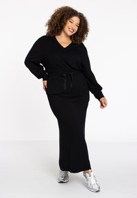 Schwarzes langärmliges Kleid mit V-Ausschnitt und Kordelzug in der Taille. Weicher Stoff, lockere Passform, getragen mit silbernen Turnschuhen.