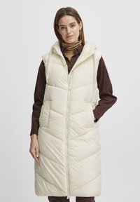 Doudoune longue beige sans manches avec une capuche, fermeture éclair à l'avant et poches latérales. Présente un matelassage en chevrons et une texture lisse et douce.