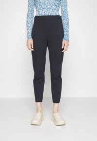 Kvinna i tapered svarta byxor, långärmad topp med blå och vit mönstring, och beigefärgade slip-on skor med texturerade sulor, står på ett vitt golv.