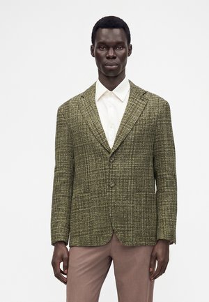 Man in een groene getextureerde blazer, wit overhemd met knopen en lichtbruine broek, staand tegen een effen witte achtergrond.