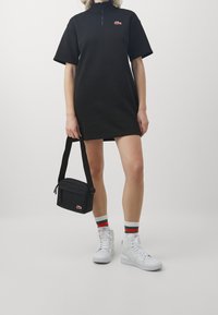 Czarna krótka sukienka na zamek błyskawiczny z logo Lacoste, w połączeniu z białymi tenisówkami sięgającymi do kostek, czarną torebką na ramię i paskowanymi skarpetkami.