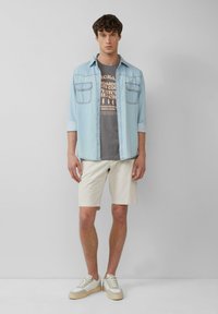 Lichtblauw denim shirt over een grijs graphic t-shirt, beige shorts en lichte sneakers. Het shirt heeft twee borstzakken; het t-shirt toont tekst.