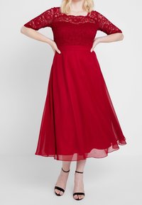 Punakol dresses dengan bodice renda dan lengan pendek, rok mengalir terbuat dari kain ringan, serta sandal hitam dengan tali. Tekstur yang halus dan desain yang elegan.