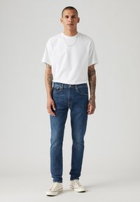 Weißes T-Shirt, blaue Slim-Fit-Jeans und weiße Sneaker. Das Model hat Tätowierungen und eine kurze Frisur und steht vor einem schlichten Hintergrund.