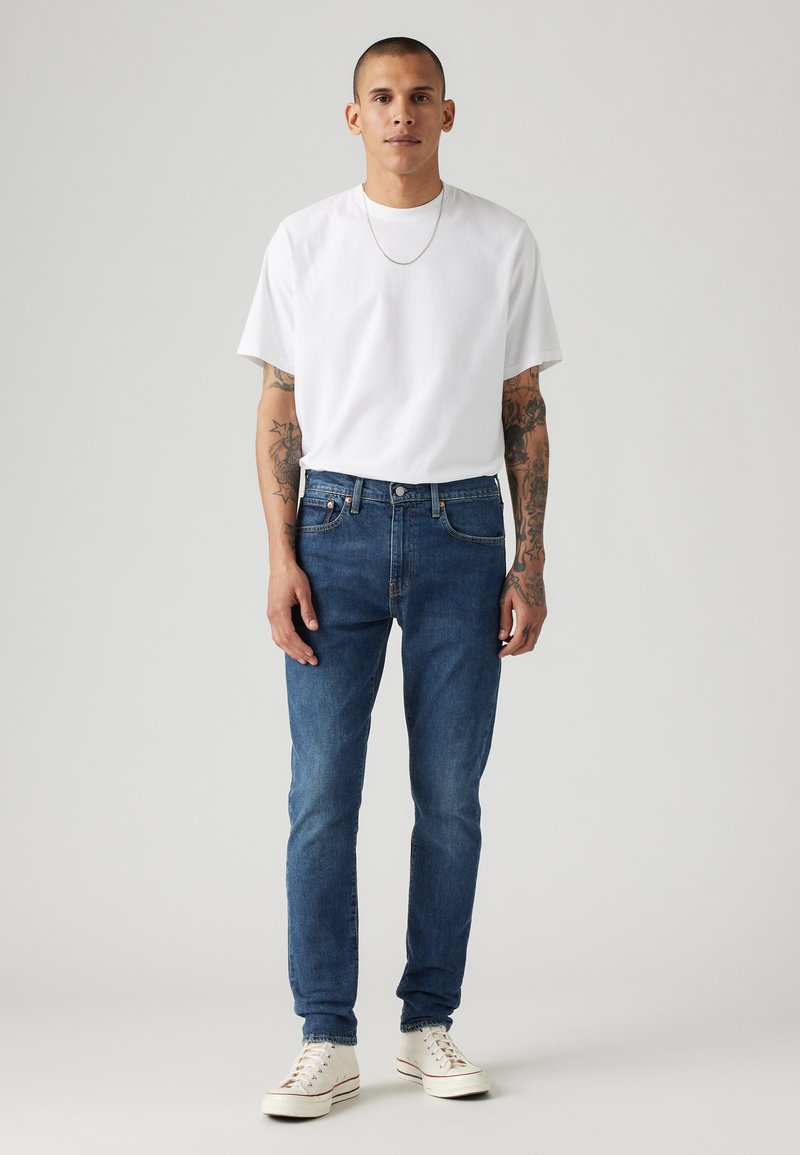 Witte T-shirt, blauwe slim-fit jeans en witte sneakers. Model heeft tatoeages en een kort kapsel, staand tegen een effen achtergrond.