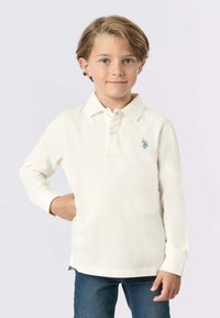 Polo shirt a maniche lunghe di colore crema con colletto, due bottoni e un logo azzurro chiaro sul petto, abbinato a jeans di denim.