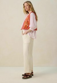 Ensemble en mélange de coton comprenant un gilet brodé orange porté sur une blouse rose clair, associé à un pantalon droit crème et des sandales marron.