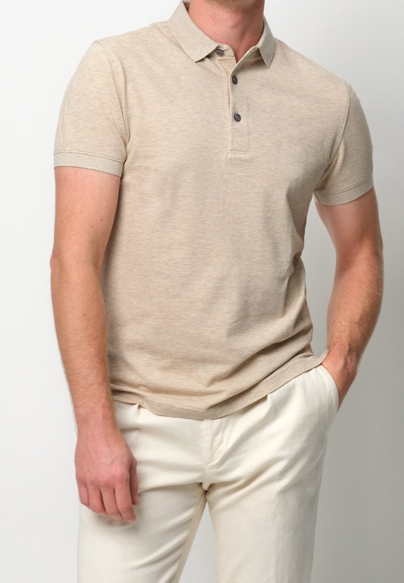 Polo beige avec col, manches courtes et trois boutons, accompagné de pantalons de couleur claire, tissu à l'apparence texturée et douce.