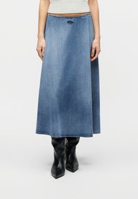 Μπλε τζιν φούστα μέσης μήκους με ελαφρά φούστα, φορεμένη με μαύρα μποτάκια με μυτερή μύτη, εμφανιζόμενη από τη μέση έως τα πόδια σε λευκό φόντο.