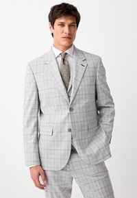 Traje gris claro con un patrón a cuadros, que presenta una chaqueta de un solo botón, solapas en pico y botones marrones. Combinado con una corbata de lunares.