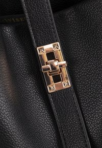 Zwarte leren band met een metalen gesp, voorzien van vierkante studs en een centrale sluiting. De textuur is gecorrigeerd en het beslag is glanzend.