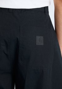 Zwarte broek van textuurstof met twee achterzakken. Een vierkante zwarte patch met een logo is zichtbaar op de rechterzak.