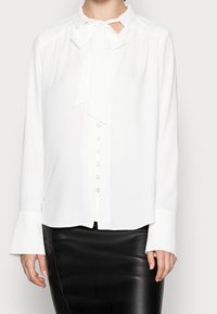 Blusa bianca con colletto a cravatta, chiusura a bottoni frontale e maniche lunghe con polsini. Tessuto morbido con una superficie liscia. Indossata con una gonna nera.