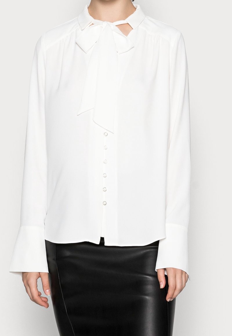 Blusa bianca con colletto a cravatta, chiusura a bottoni frontale e maniche lunghe con polsini. Tessuto morbido con una superficie liscia. Indossata con una gonna nera.