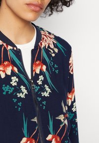 Femme portant une veste bomber bleu marine à imprimé floral rouge, blanc et vert sur un t-shirt blanc côtelé, fermeture éclair partiellement fermée.
