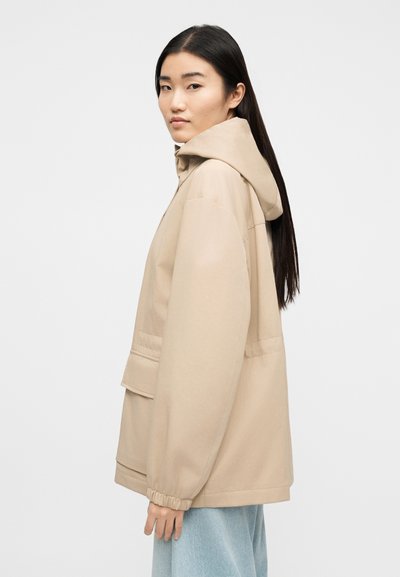 Νέα γυναίκα με ίσια μαύρα μαλλιά που φοράει μπεζ μπουφάν με κουκούλα και τσέπες, καθώς και ανοιχτό μπλε τζιν, κοιτάζοντας προς τα αριστερά σε λευκό φόντο.