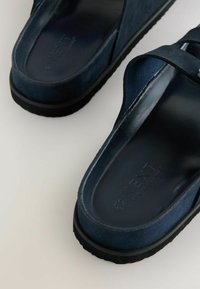 Zapatillas de ante azul marino sin cordones con una suela negra lisa y brillante, y un diseño de tiras cruzadas. El texto en la plantilla dice "NEXT."