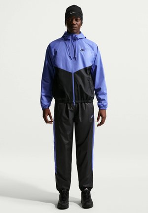 Mężczyzna ubrany w czarny i niebieski dres Nike z kurtką z kapturem, czarną czapkę i czarne sportowe buty, stoi zwrócony przodem.