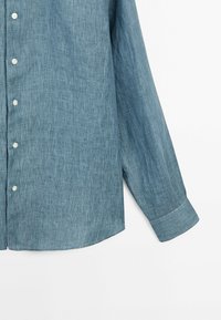 Chemise homme à manches longues boutonnée en tissu texturé bleu-gris avec des boutons blancs, montrée de la moitié du torse jusqu'au poignet sur fond blanc.