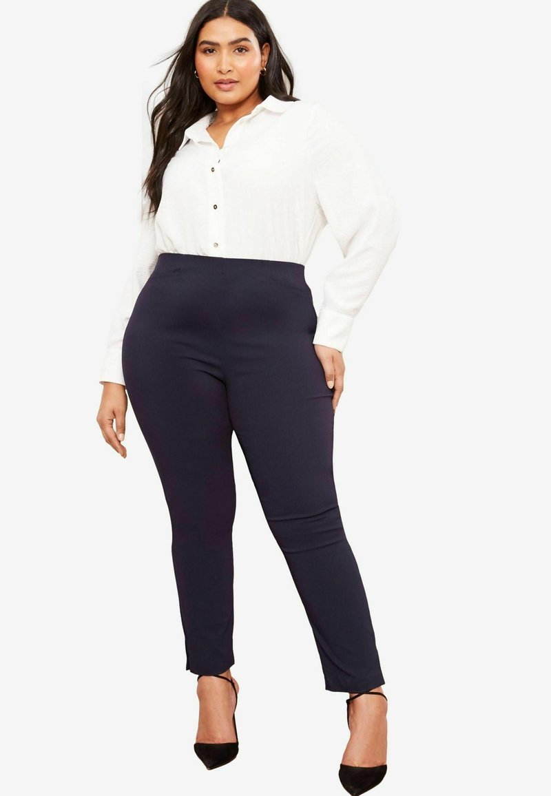Camicia bianca con bottoni e maniche lunghe abbinata a pantaloni blu scuro aderenti. I pantaloni hanno una texture liscia e una leggera svasatura alla caviglia, completati da tacchi neri.
