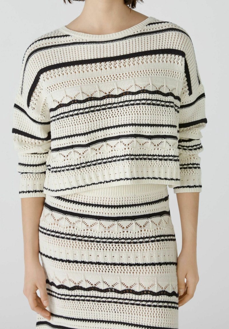 Femme portant un pull en maille crème et une jupe assortie avec des rayures horizontales noires et des motifs ajourés sur un fond uni.