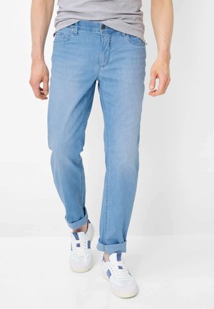 Mand iført lyseblå bukser med optrukne ben, grå T-shirt og hvide sneakers med blå detaljer, stående mod en hvid baggrund.