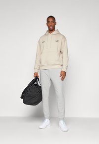 Felpa beige con tasca frontale e logo nero, abbinata a pantaloni della tuta grigi. Scarpe da ginnastica bianche e una borsa da palestra nera.