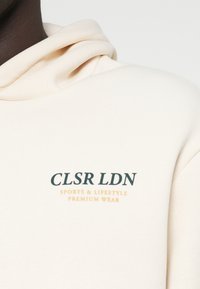 Κρέμ κάλυμμα με μαλακή υφή. Διαθέτει τσέπη καγκουρό και κεντημένο κείμενο: "CLSR LDN SPORTS & LIFESTYLE PREMIUM WEAR."