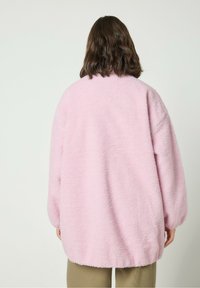 Roosa üle mõõtmete fliisjakks, millel on pehme tekstuuriga viimistlus, alusvaatega langevate õlgade ja venivate mansettidega. Tagantvaates on ümar hem.