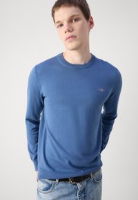 Kék crewneck pulóver hosszú ujjal, puha tapintással, bordázott mandzsettákkal és szegéllyel, a mellkason egy kis emblémával. Világoskék farmernadrággal viselve.
