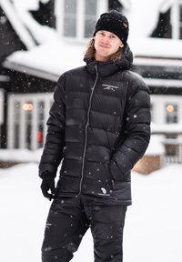 Svart isolerad jacka med dragkedja fram, puffy-design och dunfyllning, i kombination med matchande isolerade byxor, placerad i en snöig miljö.