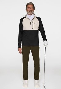 Pull-over noir et beige avec une demi-fermeture éclair, col intérieur crème, associé à un pantalon vert olive et des baskets blanches, tenant un club de golf.