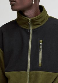 Chaqueta de forro polar con cremallera en verde y negro. Presenta un alto cuello, bolsillo lateral con cremallera, textura suave y bloques de color contrastantes.