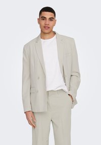 Blazer gris claro de doble botonadura en un corte ajustado, hecho de tela suave. Combinado con una camiseta blanca sencilla y pantalones a juego.
