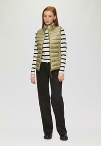 Helles olivgrünes Daunenweste mit hohem Kragen, kombiniert mit einem schwarz-weiß gestreiften Langarmshirt und schwarzen, weiten Hosen.