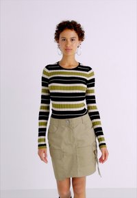 Csíkos bordás pulóver zöld, fekete és fehér színekben, bézs cargo szoknyával párosítva, amely zsebekkel és csomó részlettel rendelkezik.