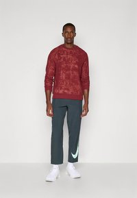 Felpa rossa con texture e polsini a coste, abbinata a pantaloni grigi scuri a zampa stretta con logo Nike verde chiaro. Indossata con sneakers bianche.