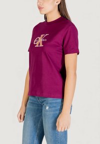 Camiseta de algodón color granate con mangas cortas, que presenta un logo dorado de "Calvin Klein" y un diseño de cuello redondo, combinada con jeans azules desgastados.