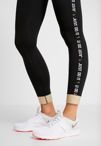 Nohy v černých legínách s textem "Just Do It" a zlatými manžetami, spárované s bílými teniskami Nike se třpytivými logy a růžovými podrážkami.