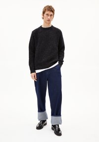 Schwarzer gerippter Strickpullover mit weißem Unterhemd, kombiniert mit weiten blauen Hosen mit umgeschlagenen Säumen und schwarzen Schuhen. Minimalistisches Design.