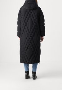 Lange zwarte pufferjas met diamanten quilt, een capuchon en oversized mouwen. Gekozen over een blauwe spijkerbroek en zwarte enkellaarsjes met hak.