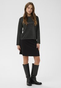 Žena v lesklém černém topu s dlouhým rukávem, černé sukni po kolena a černých vysokých botách, stojící na neutrálním pozadí.