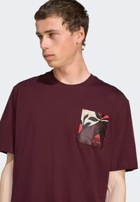 Bordeaux katoenen t-shirt met een bloemenpatroon op de zak in rood en zwart. Korte mouwen, ronde halsontwerp.