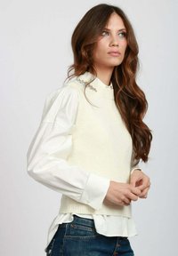 Gilet in maglia color crema con scollo a costine indossato sopra una camicia bianca con bottoni. Il look è completato da maniche lunghe e jeans in denim.