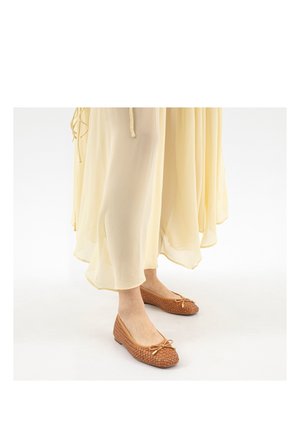 Brązowe, plecione baleriny z kokardką noszone z długą, lejącą się jasnżółtą sukienką na jednolitym białym tle.