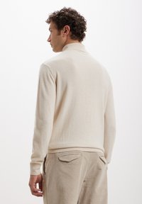 Cremfarbener Strickpullover mit einer weichen Textur und Kragen. Die Hinteransicht zeigt ein figurbetontes Design. Getragen mit hellbeigen Cordhosen.